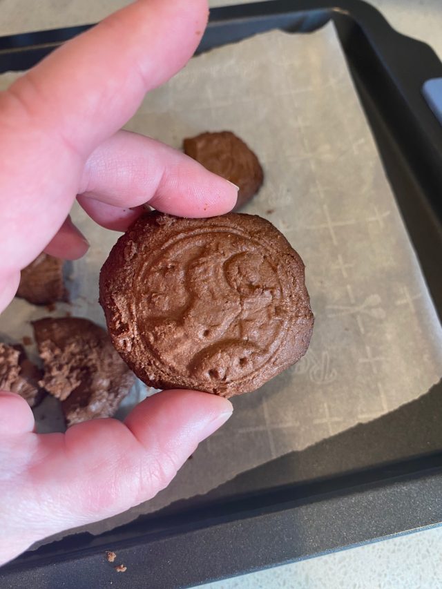 A hand holding a homemade mooncake with a stamped design, visibly overbaked and cracked, resting above a parchment-lined baking sheet.