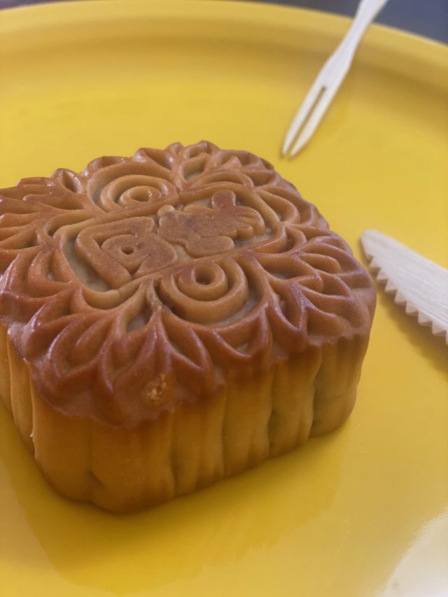A traditional mooncake with an intricate floral pattern sits on a bright yellow plate, with a small plastic fork nearby.