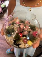 A finished terrarium - a small fishbowl-sized glass container with a pink plant surrounded by small mossy plants and 3D printed items like flamingoes and mushrooms.